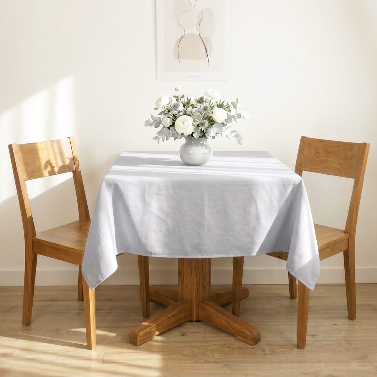 Table en bois deux couverts contre un mur, la table est dressée avec la nappe métis grise, au centre un vase posé sur la table avec un bouquet de fleurs. Décor minimaliste et très lumineux.