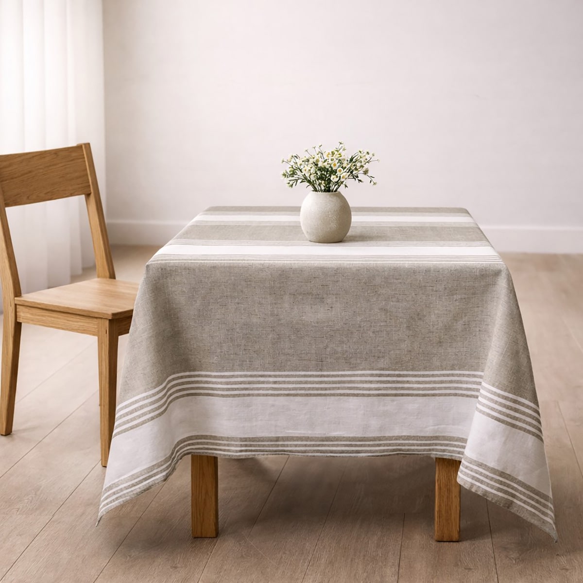 Table carrée recouverte d’une nappe beige à rayures de couleur blanche, avec un petit vase de fleurs au centre, dans une pièce lumineuse au style minimaliste, avec une chaise en bois à côté.
