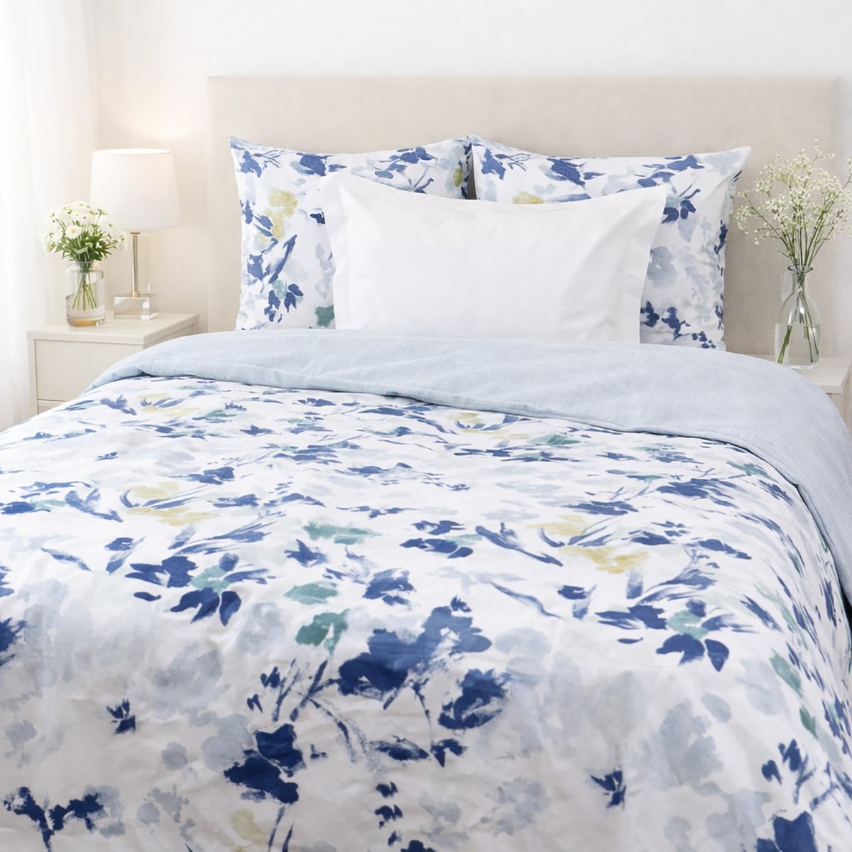 Lit double avec parure de lit blanche à fleurs bleues et un revers uni strié bleu très léger, accompagné de deux oreillers assortis, dans une chambre lumineuse avec table de chevet en bois, lampe blanche et petite plante verte.