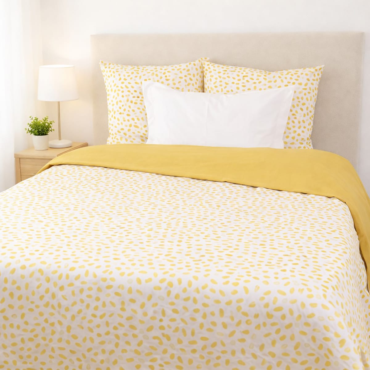 Lit double avec parure de lit blanche à motifs jaunes et housse de couette unie jaune, accompagné de deux oreillers assortis, dans une chambre lumineuse avec table de chevet en bois, lampe blanche et petite plante verte.